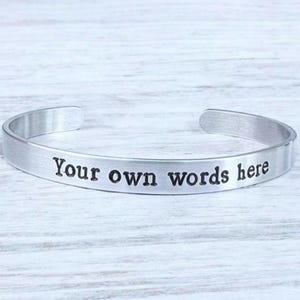 Puede incluir: Una pulsera de puño plateada con las palabras "Your own words here" grabadas. La pulsera abierta se encuentra sobre una superficie clara y texturizada.