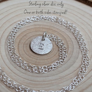 Könnte beinhalten: Eine Halskette aus Sterlingsilber mit einem runden Scheibenanhänger mit strukturierter Oberfläche und dem eingestanzten Buchstaben E in der Mitte. Der Text Sterling silver disc only One or both sides stamped! ist über dem Anhänger zu sehen.