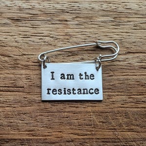 Op de afbeelding: Zilveren veiligheidsspeld met een rechthoekige hanger met de tekst "I am the resistance" in een vetgedrukte lettertype. De speld is geplaatst tegen een getextureerde, houten achtergrond.
