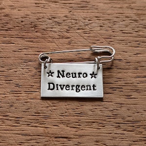 Peut inclure: Épingle de sûreté argentée avec une étiquette rectangulaire. L'étiquette porte les mots "Neuro Divergent" en caractères gras, avec une étoile de chaque côté du mot "Neuro". L'épingle est posée sur une surface en bois.