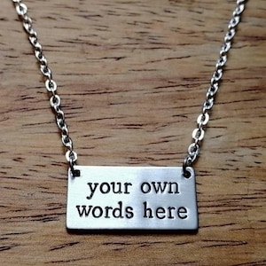May include: Silver necklace featuring a rectangular pendant engraved with the words "your own words here". The chain is a delicate link design. The pendant hangs against a wooden background, creating a simple, elegant aesthetic.