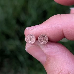 May include: A pair of small, circular rose gold stud earrings, each featuring a textured, hammered surface and the capital letter J embossed in the center. The earrings have a metallic finish and a distinct, uneven, rugged appearance.