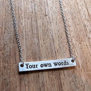 May include: A silver-toned metal bar necklace featuring a delicate chain and a rectangular pendant. The pendant is engraved with the black text "Your own words..." in a simple, sans-serif font, centered horizontally on the brushed metal surface.