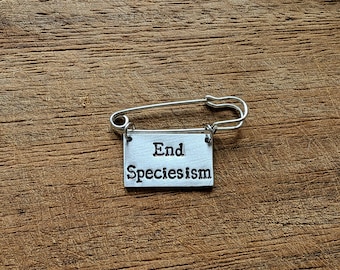 May include: A silver safety pin with a rectangular tag that reads "End Speciesism" in a bold, sans-serif font. The pin is set against a textured, brown wooden surface.