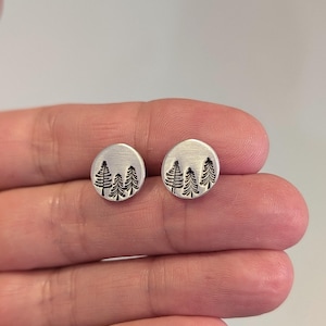 May include: A pair of silver stud earrings. Each earring has a circular design with an etched image of three stylized evergreen trees. The earrings are small and minimalist, suitable for everyday wear. The earrings are shown against a neutral background.