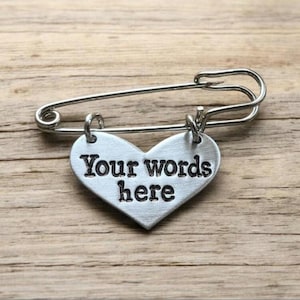 May include: A silver safety pin with a heart-shaped charm. The charm is inscribed with the words "Your words here". The pin rests on a wooden surface.