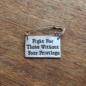 May include: A silver safety pin with a rectangular charm. The charm reads: "Fight For Those Without Your Privilege". The pin is set against a wooden background.