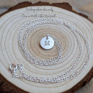 Peut inclure: Un collier en argent sterling avec un petit pendentif disque circulaire estampillé de la lettre M. La chaîne est de style maillons fins en argent avec un fermoir à ressort rond. Le texte "Sterling silver disc only One or both sides stamped!" est visible.