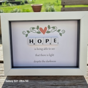 May include: A white frame with a black mount and a white background. The word "HOPE" is spelled out with Scrabble tiles. The tiles are arranged in a line with a small red heart above them. The quote "Hope is being able to see that there is light despite the darkness" is written below the tiles.