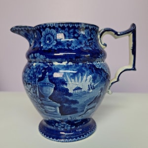 May include: A blue and white ceramic pitcher with a floral pattern on the top and a landscape scene on the body. The pitcher has a handle and a spout. The landscape scene includes trees, a building, and figures.