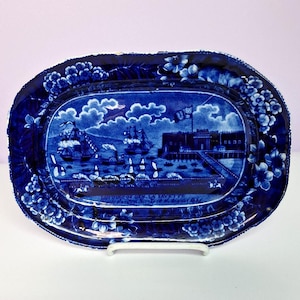 May include: An antique blue and white ceramic platter with a nautical scene. The oval-shaped platter features a detailed illustration of ships, buildings, and a flag. Floral patterns adorn the rim. The platter is displayed on a white stand.