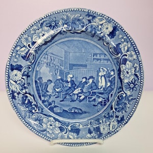 May include: Blue and white ceramic plate with a detailed illustration of a gathering scene. The central image is surrounded by a floral border. The plate is supported by a white stand, showcasing its decorative design.