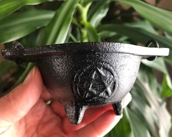 May include: A small black cast iron cauldron with three legs and a pentagram design. The cauldron has two small handles on the sides.