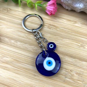 May include: A blue glass Evil Eye keyring with a silver metal ring and chain. The keyring features a large, round charm and a smaller charm, both with a white and blue eye design. The background is a light brown wooden surface.