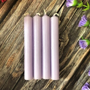 May include: Four lavender-coloured candles, each with a white wick, are arranged side-by-side on a wooden surface.