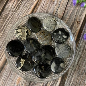 Peut inclure: Une collection de pierres circulaires lisses et polies dans des tons de noir, gris et crème, disposées sur une assiette en verre transparent texturé. Les pierres présentent des motifs naturels variés, incluant des surfaces noires unies, des textures grises marbrées et des veines crème complexes.