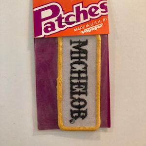 May include: Iron-on patch with a white background and yellow border. The patch has black text that reads "MICHENER" with a registered trademark symbol.
