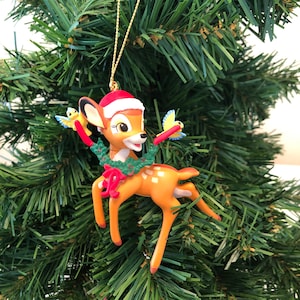 Peut inclure: Une décoration de Noël représentant un cerf de dessin animé portant un chapeau de Père Noël et tenant une couronne. Deux petits oiseaux jaunes sont perchés sur les bois du cerf.