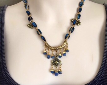 Puede incluir: Un collar azul y dorado con un colgante que presenta un diseño geométrico y piedras azules. El collar está hecho con cuentas ovaladas azules y detalles dorados.