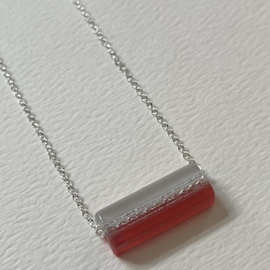 May include: A silver chain necklace with a rectangular pendant. The pendant is divided into three sections: a top gray section, a middle clear section, and a bottom red section. The necklace is displayed on a white surface.