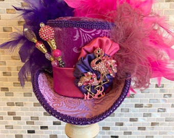 May include: A decorative purple top hat with pink and purple feathers. The hat features a pink satin band, a decorative flamingo brooch, and jeweled embellishments. The hat is displayed on a white stand.