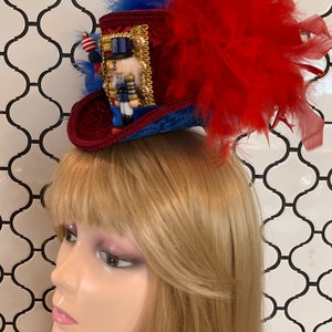 May include: A festive miniature top hat in red, blue, and gold, adorned with a nutcracker figurine and red feathers. The hat features a velvet texture and is embellished with decorative trim and beads, perfect for holiday celebrations.