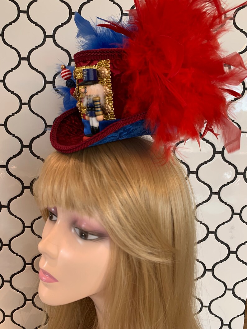 May include: A festive miniature top hat in red, blue, and gold, adorned with a nutcracker figurine and red feathers. The hat features a velvet texture and is embellished with decorative trim and beads, perfect for holiday celebrations.