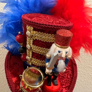 May include: A red, white, and blue decorative hat with a miniature nutcracker soldier and drum. The hat features red feathers, gold trim, and a glittery red top. The nutcracker is in a blue and red uniform, holding a silver sword.