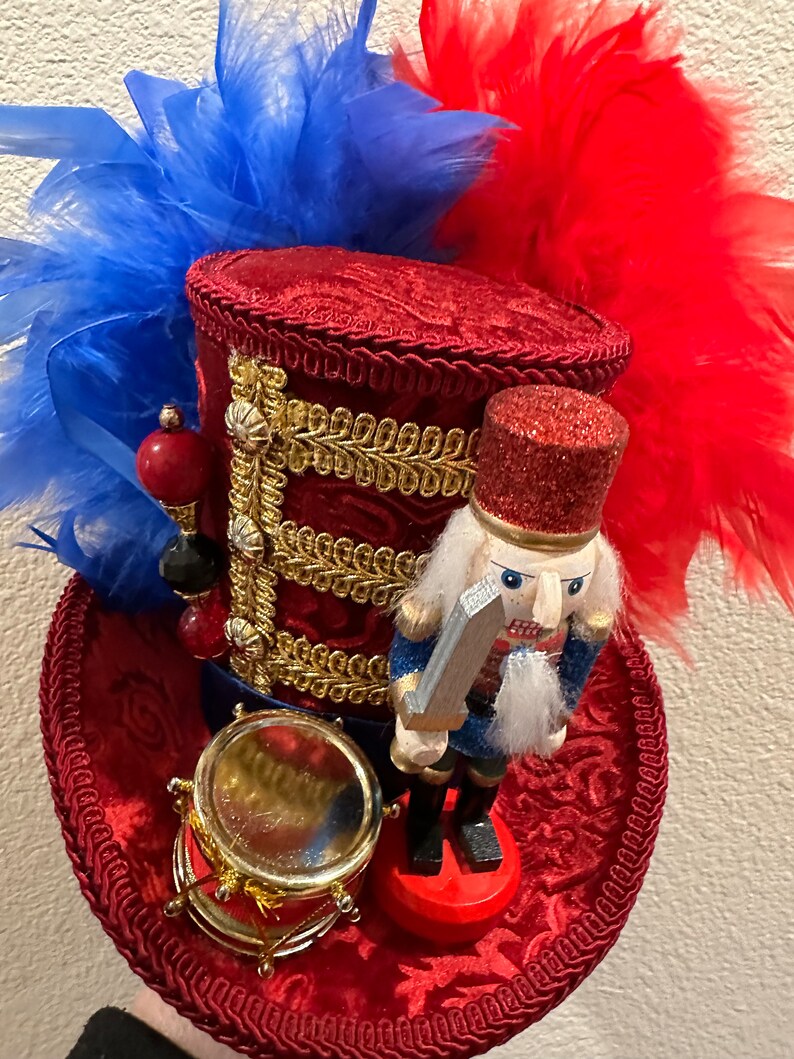 May include: A red, white, and blue decorative hat with a miniature nutcracker soldier and drum. The hat features red feathers, gold trim, and a glittery red top. The nutcracker is in a blue and red uniform, holding a silver sword.