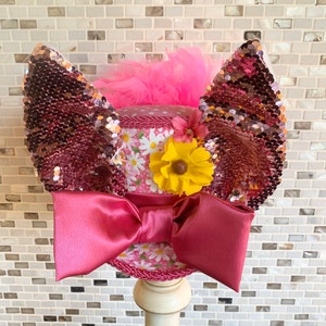 May include: A decorative hat with a daisy print, a large pink bow, and sequined ear-like accents. The hat features a yellow flower and pink feathers. The hat is displayed on a white stand.