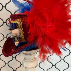 May include: A decorative red and blue hat with a nutcracker figurine. The hat features a red velvet crown, blue trim, and a large cluster of red feathers. A small nutcracker soldier is attached to the hat, with gold accents and a small American flag.
