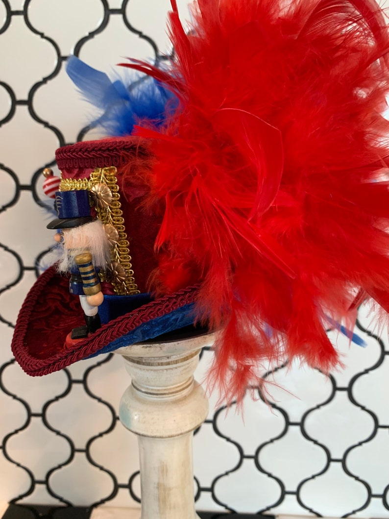 May include: A decorative red and blue hat with a nutcracker figurine. The hat features a red velvet crown, blue trim, and a large cluster of red feathers. A small nutcracker soldier is attached to the hat, with gold accents and a small American flag.