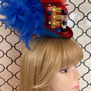 May include: A miniature top hat with red, blue, and gold accents, adorned with blue and red feathers. The hat features a decorative gold trim, a small nutcracker figurine, and red and white beads. The hat is designed to be worn.