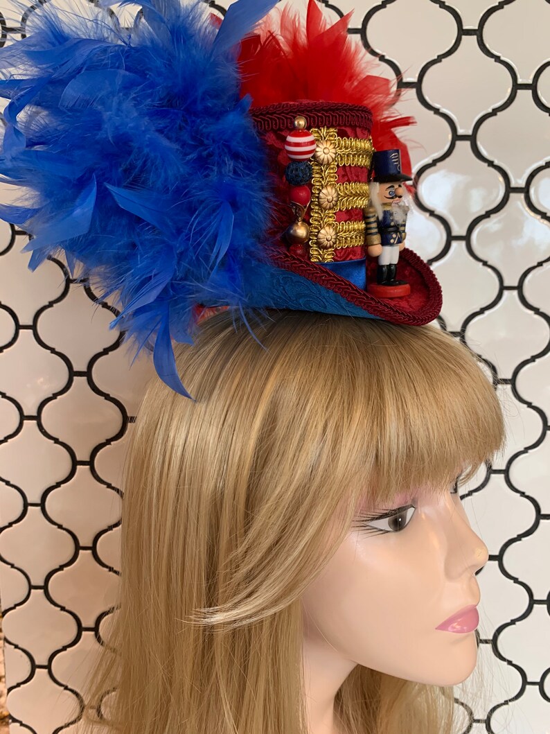 May include: A miniature top hat with red, blue, and gold accents, adorned with blue and red feathers. The hat features a decorative gold trim, a small nutcracker figurine, and red and white beads. The hat is designed to be worn.