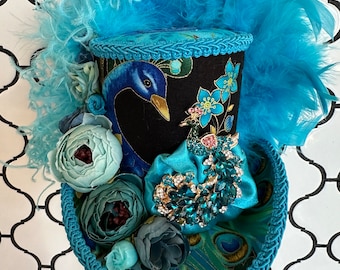 May include: A decorative top hat featuring a peacock design. The hat has a black base with a peacock illustration and blue floral accents. It is adorned with blue feathers, fabric flowers, and a jeweled embellishment. The brim is trimmed with a blue rope.