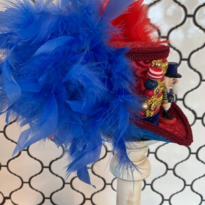 May include: A decorative hat featuring a red and blue color scheme, adorned with blue feathers. The hat has a red brim and a detailed design, with a miniature nutcracker figure on the side. The hat is displayed on a white pedestal.