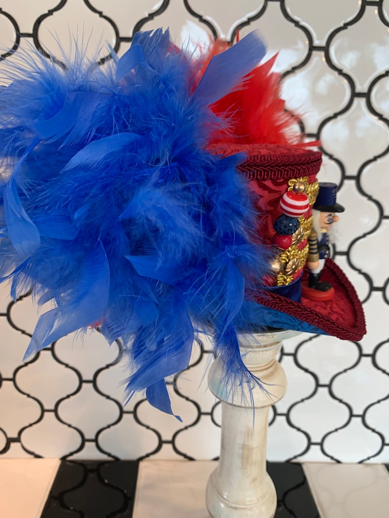 May include: A decorative hat featuring a red and blue color scheme, adorned with blue feathers. The hat has a red brim and a detailed design, with a miniature nutcracker figure on the side. The hat is displayed on a white pedestal.