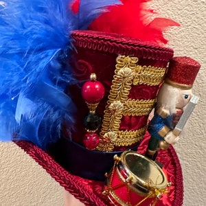 May include: A decorative red top hat with blue and red feathers. The hat features gold trim, a small drum, and a nutcracker figurine. The hat is embellished with red, black, and gold beads.