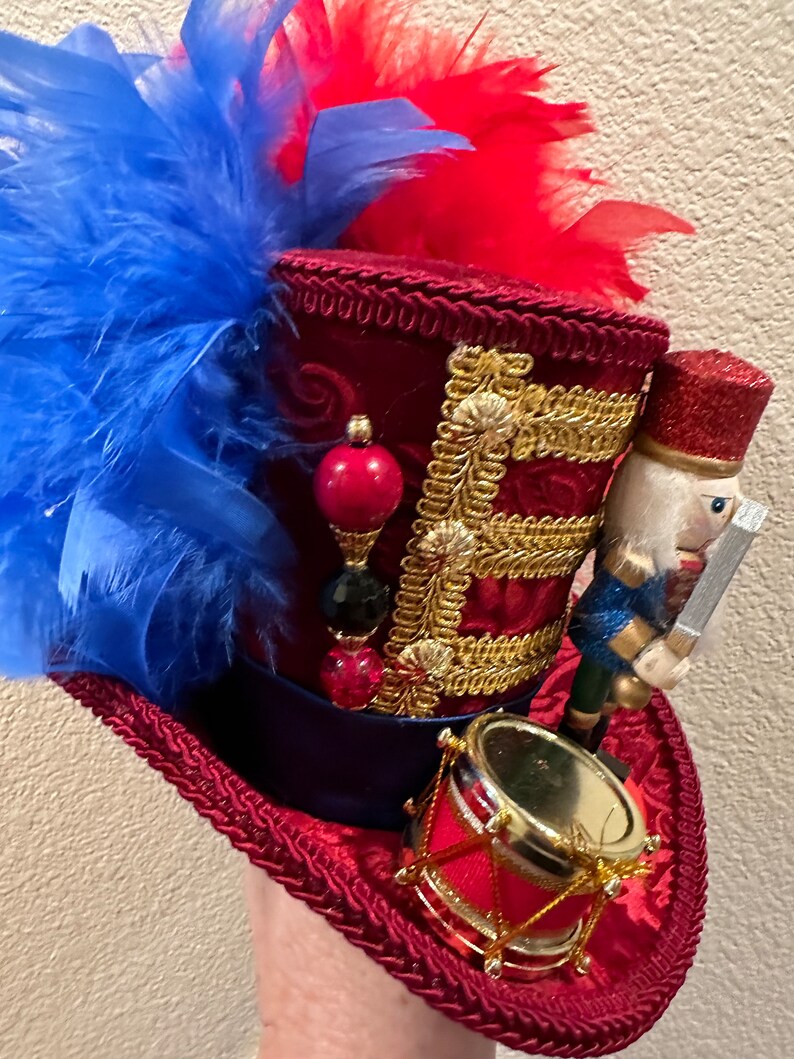May include: A decorative red top hat with blue and red feathers. The hat features gold trim, a small drum, and a nutcracker figurine. The hat is embellished with red, black, and gold beads.