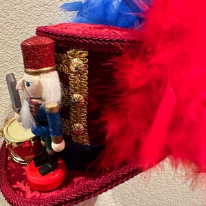 May include: A red velvet top hat with gold trim and a red, white, and blue nutcracker soldier figurine. The hat is adorned with red feathers and blue accents. A small drum is next to the nutcracker.