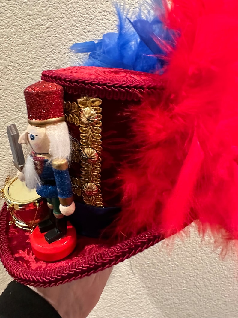May include: A red velvet top hat with gold trim and a red, white, and blue nutcracker soldier figurine. The hat is adorned with red feathers and blue accents. A small drum is next to the nutcracker.