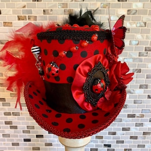 May include: A red and black ladybug-themed top hat with a polka dot pattern. The hat is adorned with ladybug embellishments, red feathers, a butterfly, and a black ribbon. The hat is displayed on a white stand.