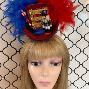 May include: A festive red, white, and blue miniature top hat with a nutcracker figurine, gold trim, and feather accents. The hat is adorned with blue and red feathers and decorative beads, perfect for holiday celebrations.