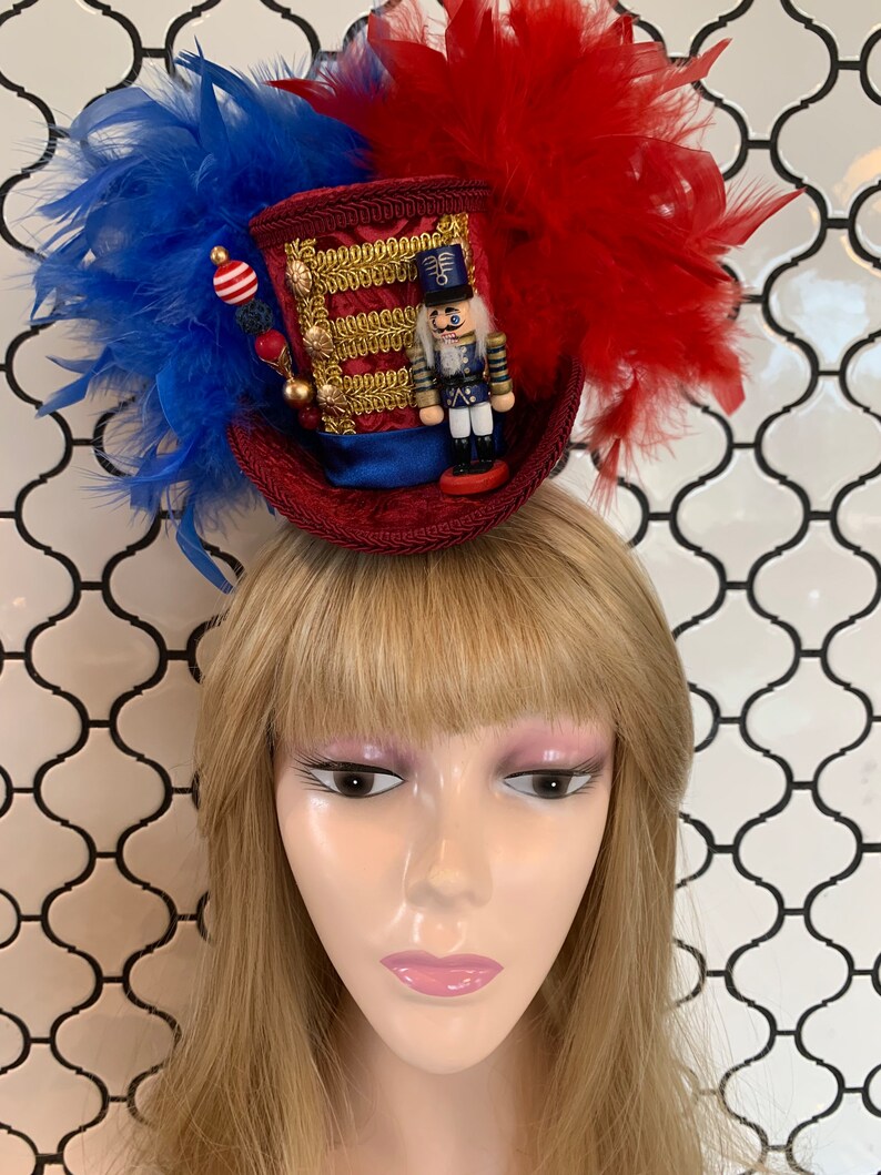 May include: A festive red, white, and blue miniature top hat with a nutcracker figurine, gold trim, and feather accents. The hat is adorned with blue and red feathers and decorative beads, perfect for holiday celebrations.