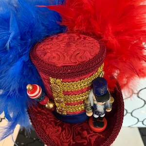 May include: A decorative red top hat with blue and red feathers. The hat features gold trim, a small nutcracker figurine, and a beaded embellishment. The hat is likely for a costume or special event.