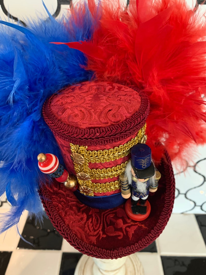 May include: A decorative red top hat with blue and red feathers. The hat features gold trim, a small nutcracker figurine, and a beaded embellishment. The hat is likely for a costume or special event.