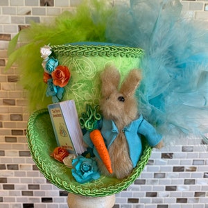 May include: A decorative Easter hat with a plush bunny wearing a blue jacket, holding a carrot and a book titled "The Tale of Peter Rabbit." The hat is green with floral accents and colorful feathers.