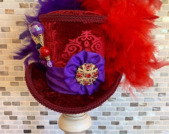 May include: A burgundy velvet top hat with a purple satin ribbon and a purple flower with a gold and red jewel. The hat is adorned with red and purple feathers and decorative beading.