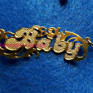May include: Gold-tone necklace with a cutout 'Baby' pendant. The pendant has a textured surface and is attached to a delicate chain.