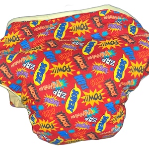 May include: A red, white, and blue fabric diaper with a superhero theme. The diaper is covered in a repeating pattern of comic book sound effects like "POW!" and "ZAP!"